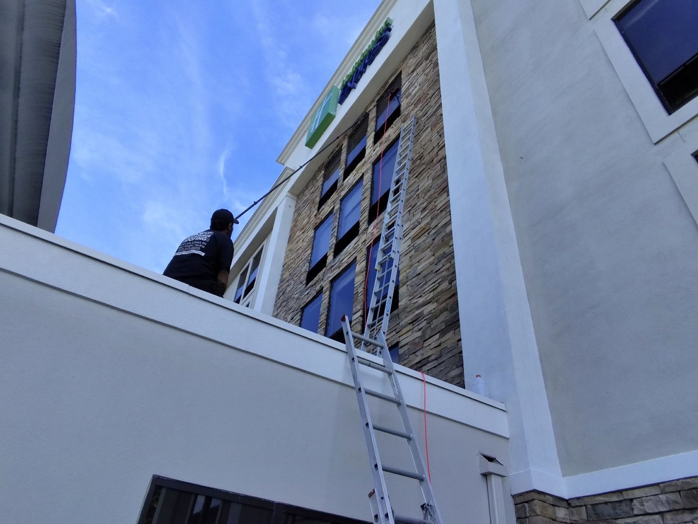 A person uses a long cleaning pole to wash windows on a stone-facade building while a ladder leans against the wall.