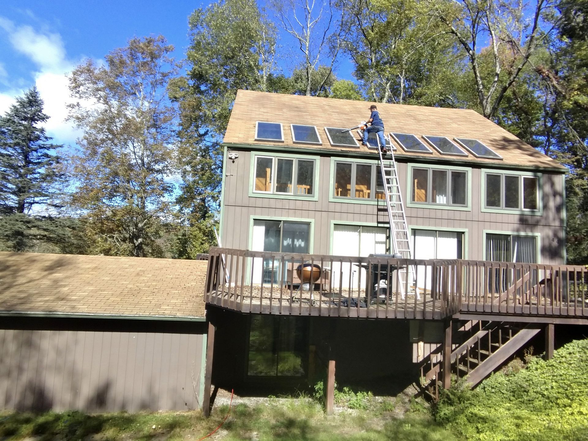 A person on a ladder installs solar panels on the shingled roof of a two-story house with a wooden deck.
