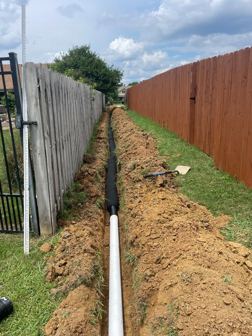 A trench dug alongside a wooden fence, containing a white pipe wrapped in black fabric.