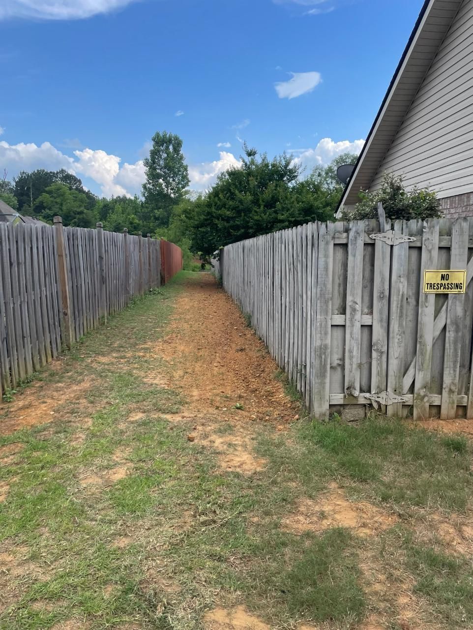 A narrow dirt path between weathered wooden fences leads towards trees under a bright blue sky.
