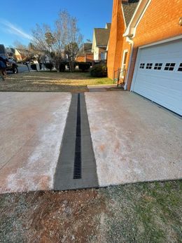 A concrete driveway with a long drain. Next to a brick house and green lawn.