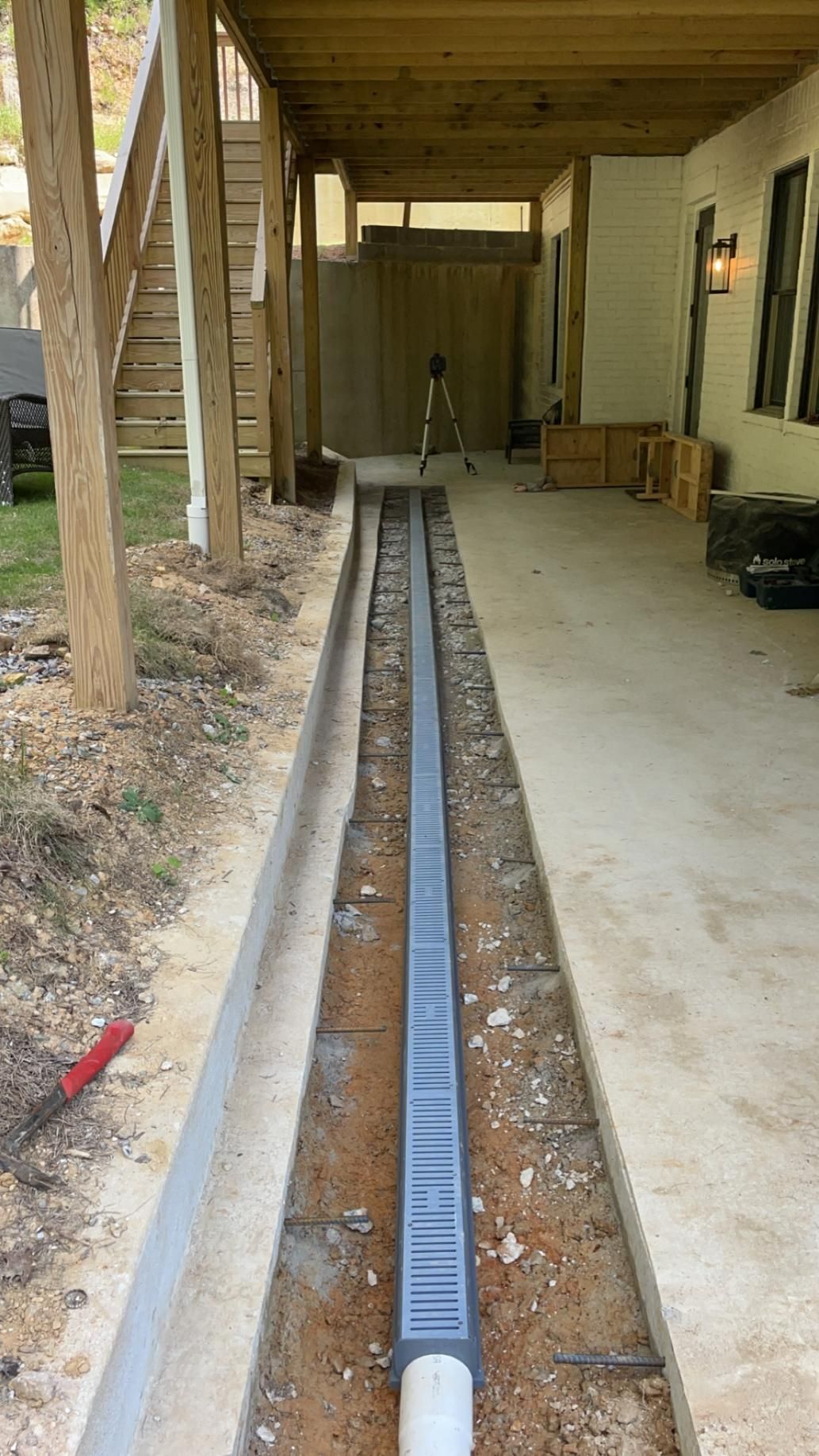 Construction site with a long trench drain installed in a concrete area; under a porch, next to a house.