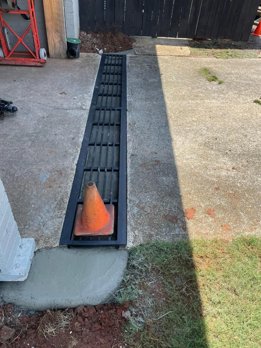 A long, black drain with a traffic cone inside sits in concrete, next to grass.