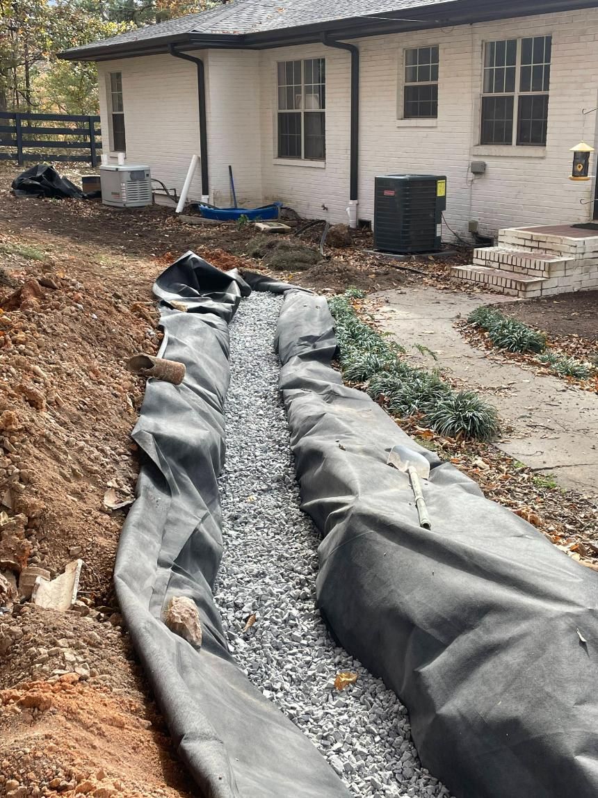 A gravel-filled trench lined with black fabric, next to a white brick house, likely for drainage.
