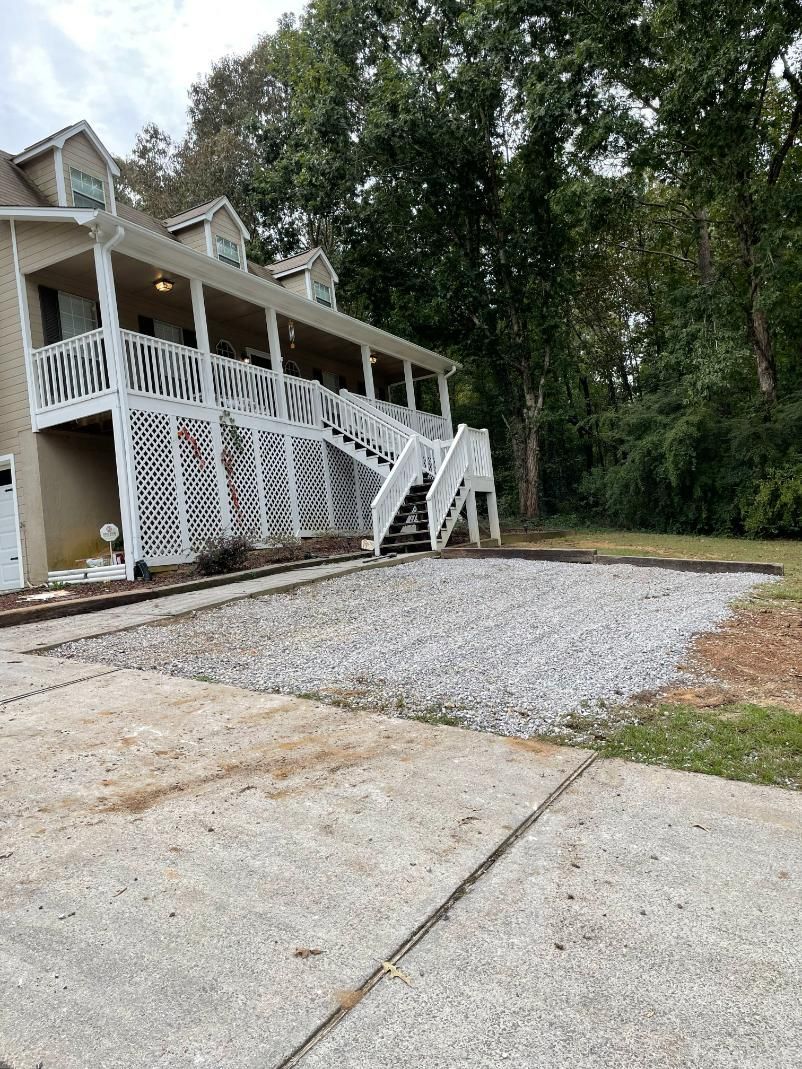 A gravel area next to a concrete driveway, leading up to a multi-level house with a white porch and stairs.