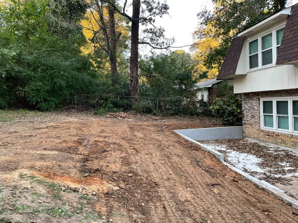 Dirt yard next to a brick house with a concrete border, with trees in the background.