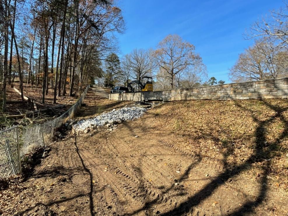 Construction site: excavator on a retaining wall, dirt, rocks, fence, bare trees, and blue sky.
