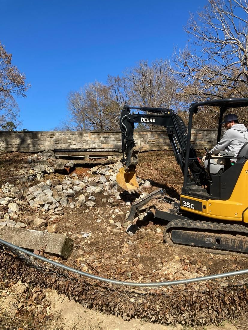 A yellow excavator with a worker is demolishing a stone wall in a sunny, outdoor setting.
