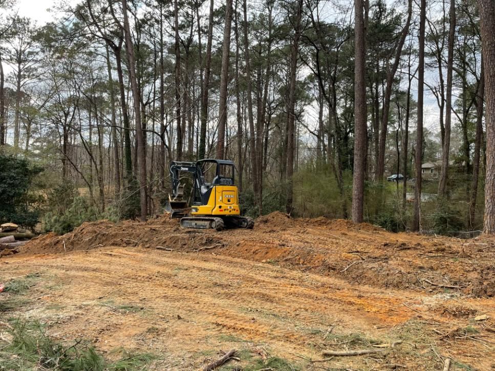 Yellow excavator clearing land in a wooded area, dirt and trees visible.