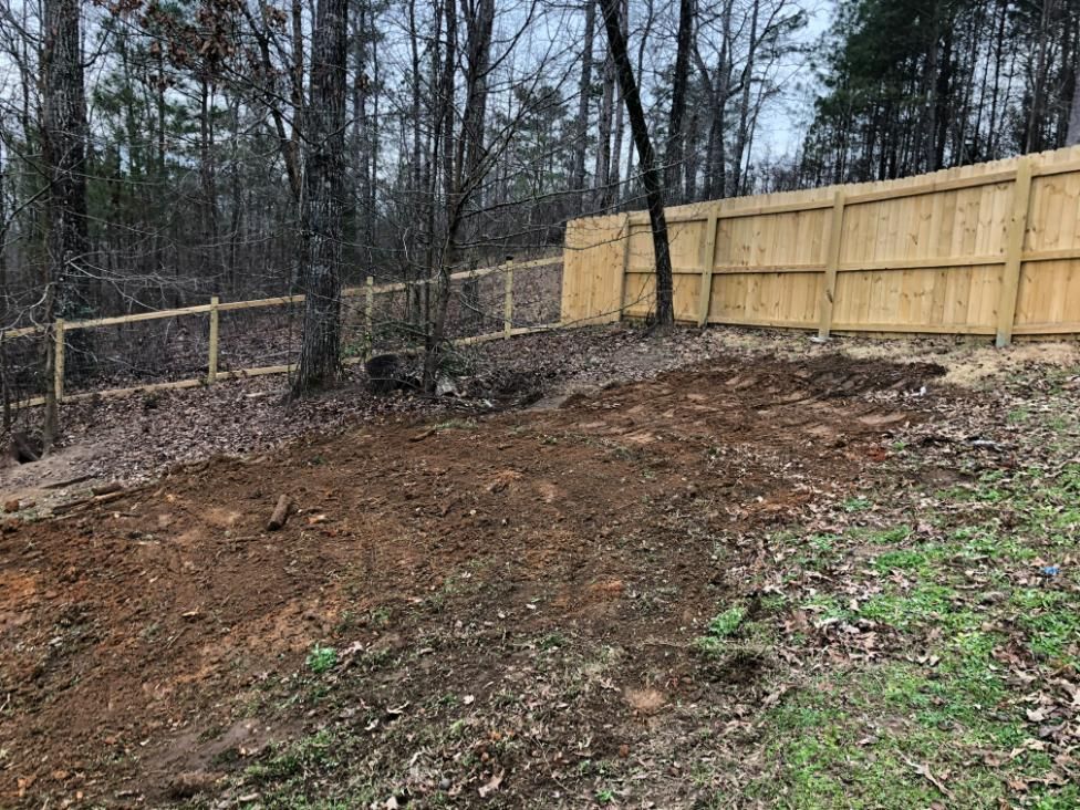 A cleared, brown dirt area on a hillside, with wooden fences and trees in the background.