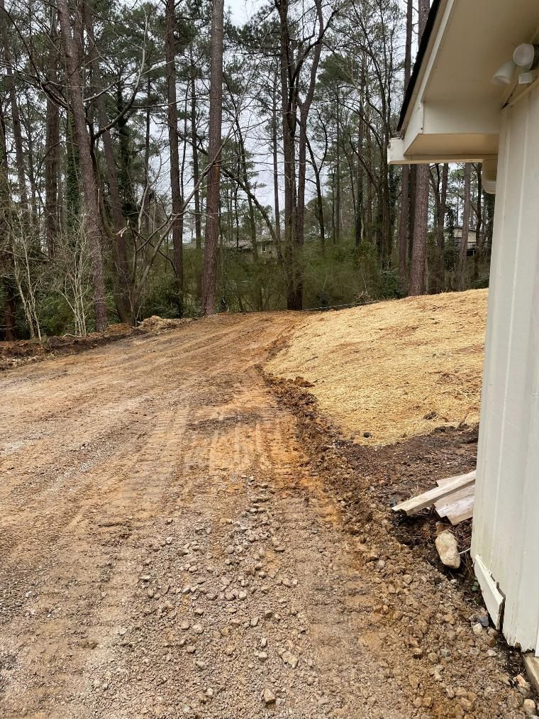 Dirt driveway with fresh grading next to a white building, trees in the background.