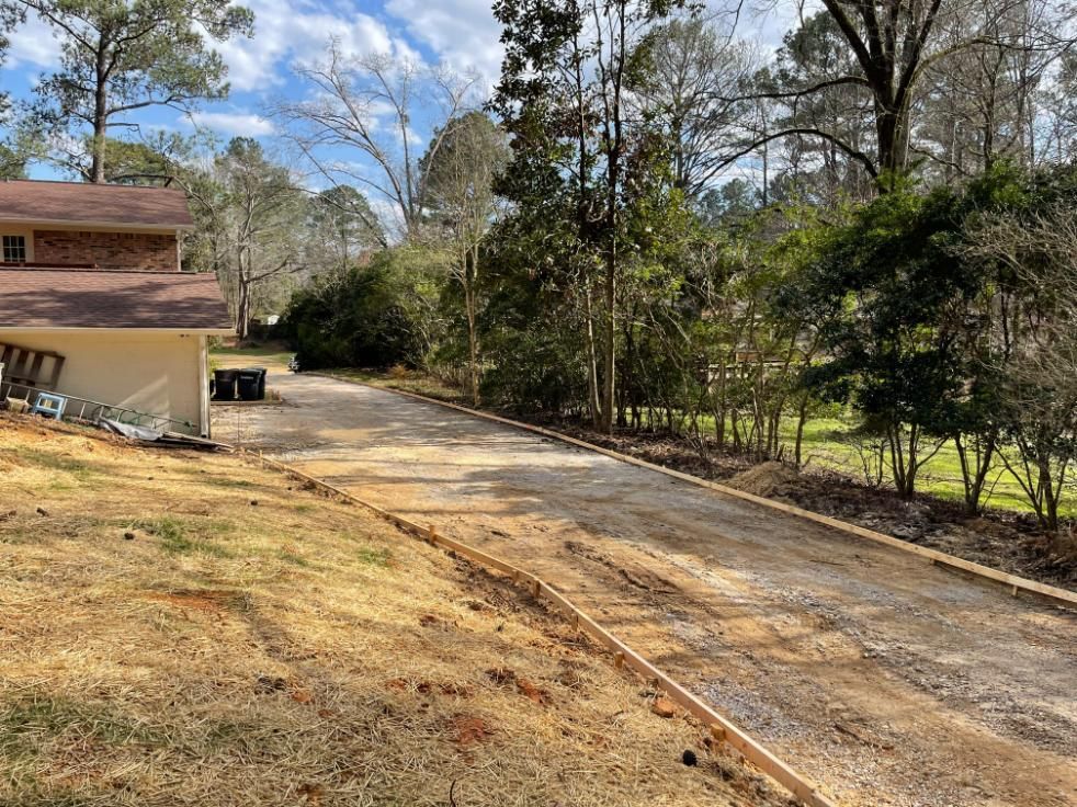 Gravel driveway next to a grassy area. Wooden borders define edges. Trees and a house are in the background.