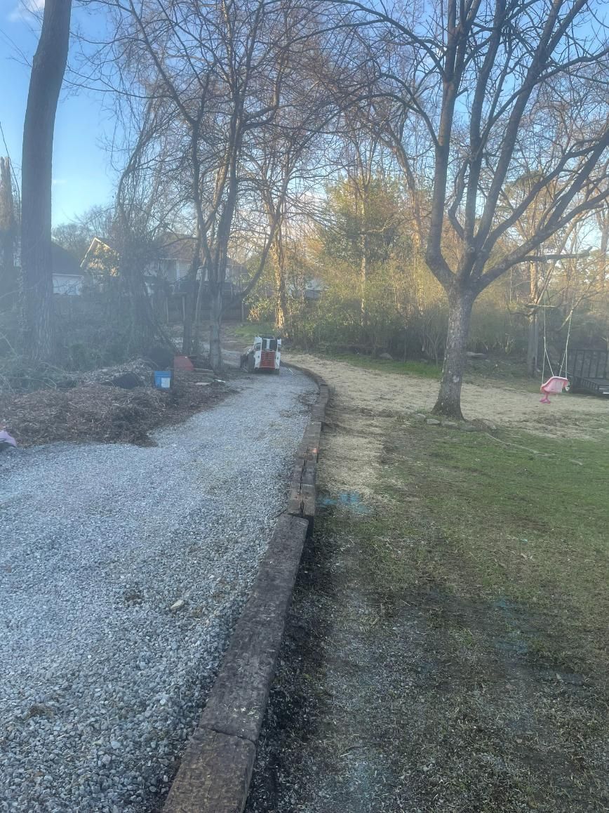 Gravel driveway curving through a yard, lined with retaining blocks, trees, and grass.