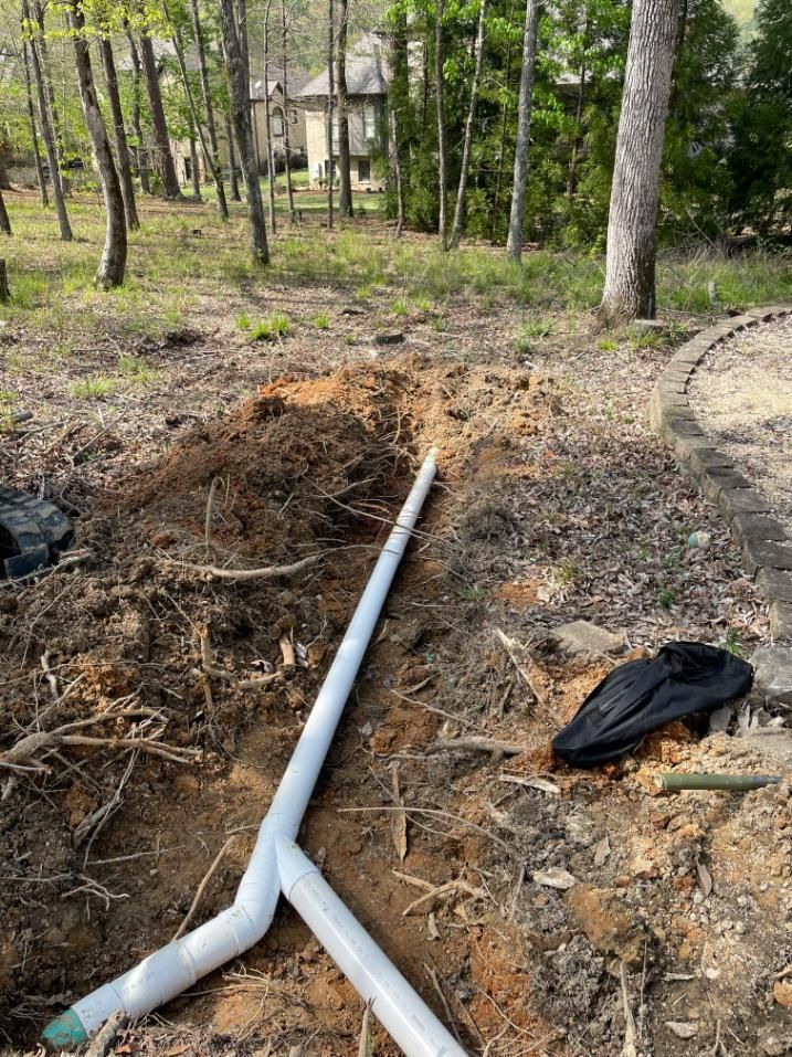 White PVC pipes in a dirt excavation, with a shoe nearby, in a wooded outdoor setting.
