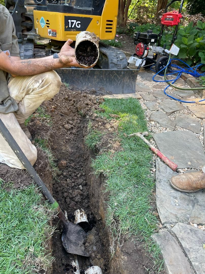 Man excavating a pipe in a trench. A yellow excavator and red engine are in the background.