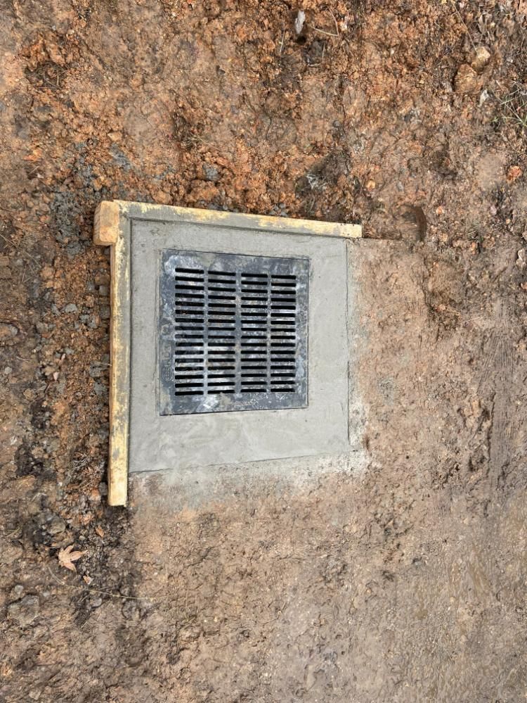 Square drainage grate in concrete, set in brown soil, surrounded by a wooden frame.