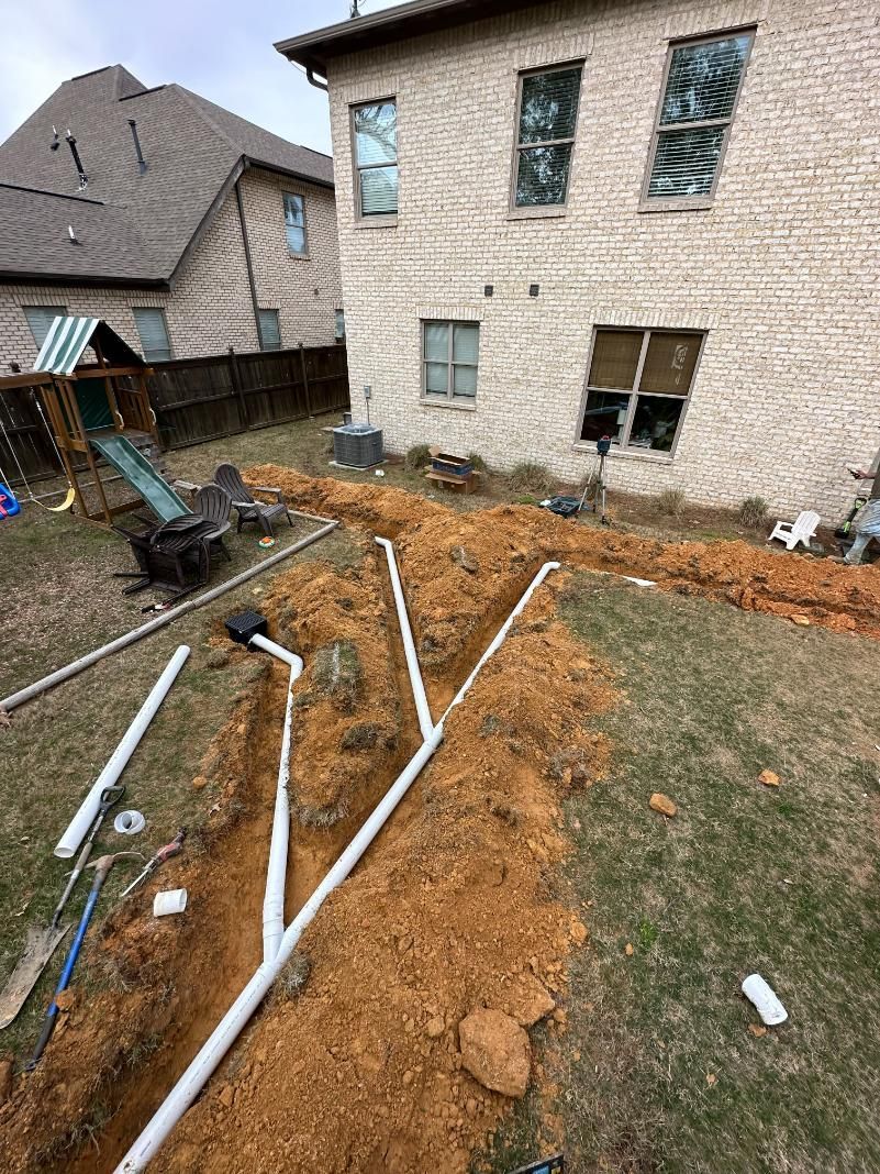 Backyard with trenches dug for white drainage pipes near a brick house, with a play set visible.
