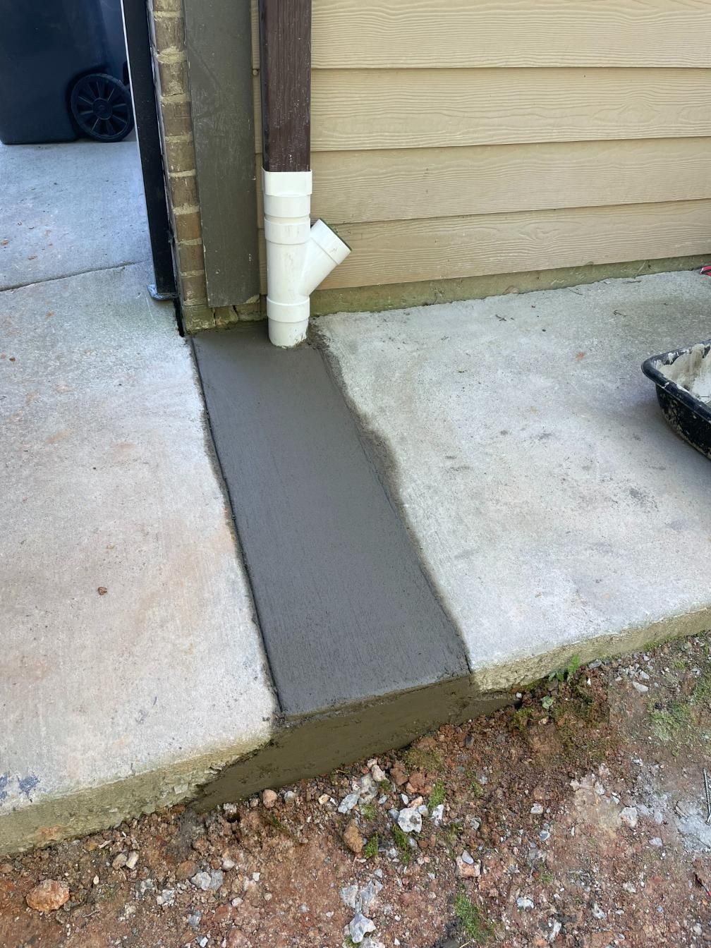 Freshly poured concrete strip next to a building, with a downspout and trash can visible.