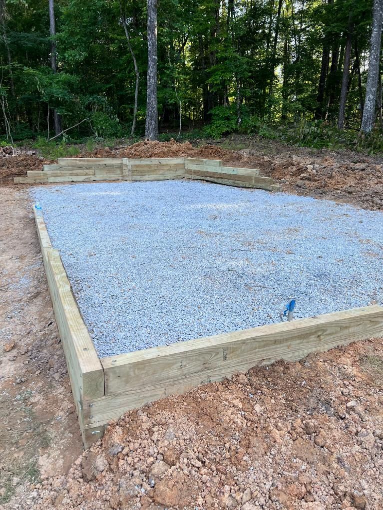 Gravel-filled rectangular platform with wooden borders, set in a dirt yard with trees in the background.