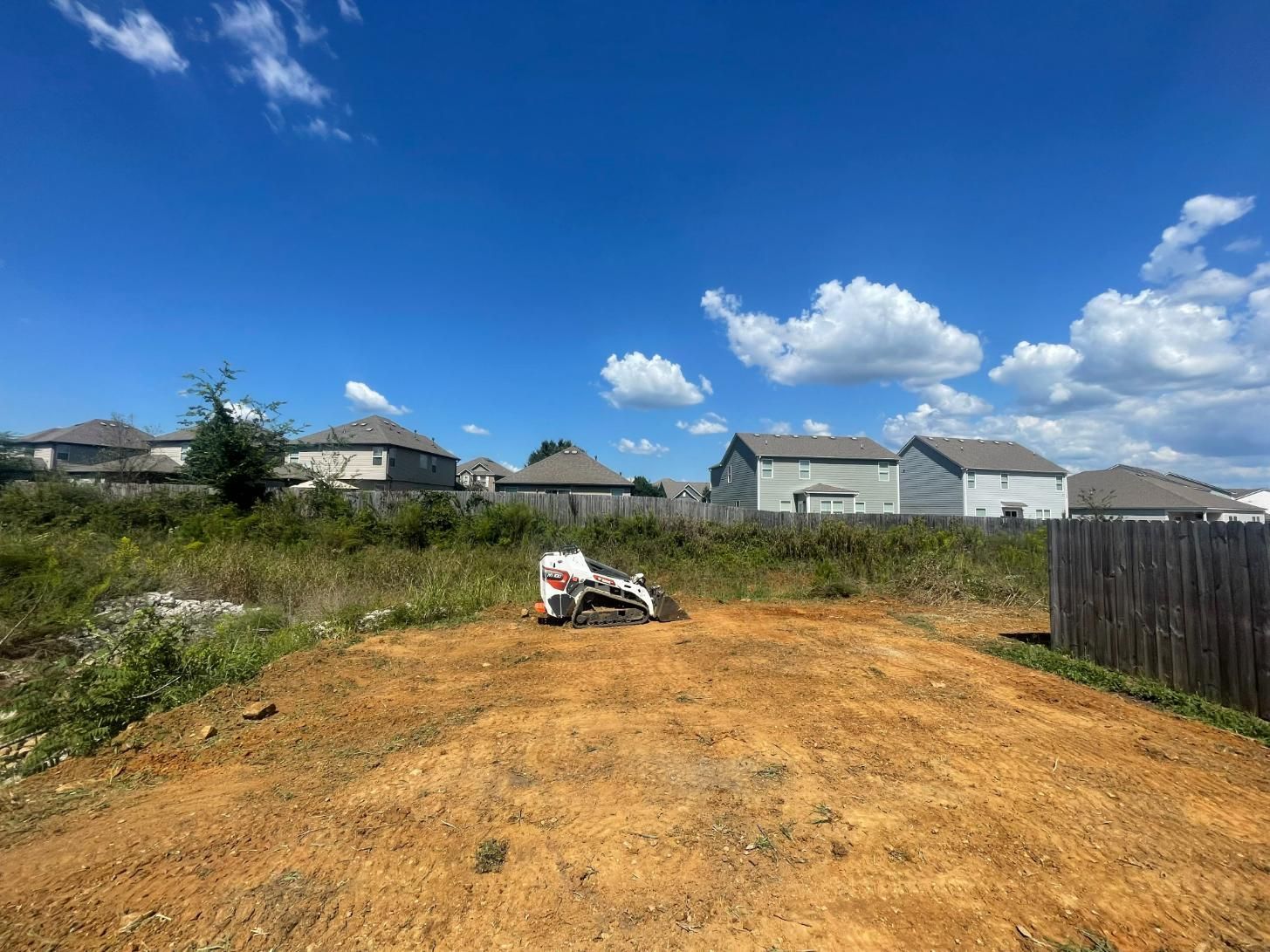 A small excavator on a dirt pile in a residential area under a blue sky with clouds.