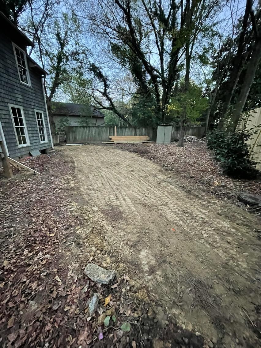 A dirt driveway next to a weathered gray house, covered in fallen leaves, leading towards trees.