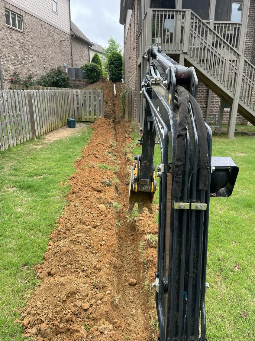 A small excavator digs a trench in a grassy backyard, near a fence and house.