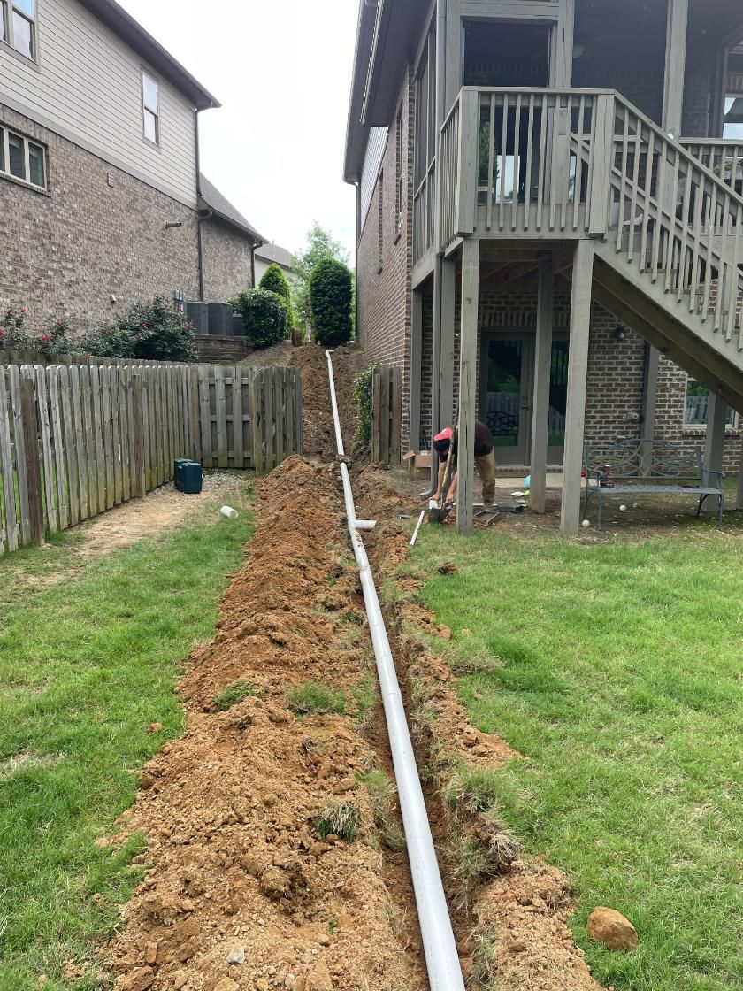 Workers installing a white pipe in a trench dug in a grassy backyard. A house and fence are visible.