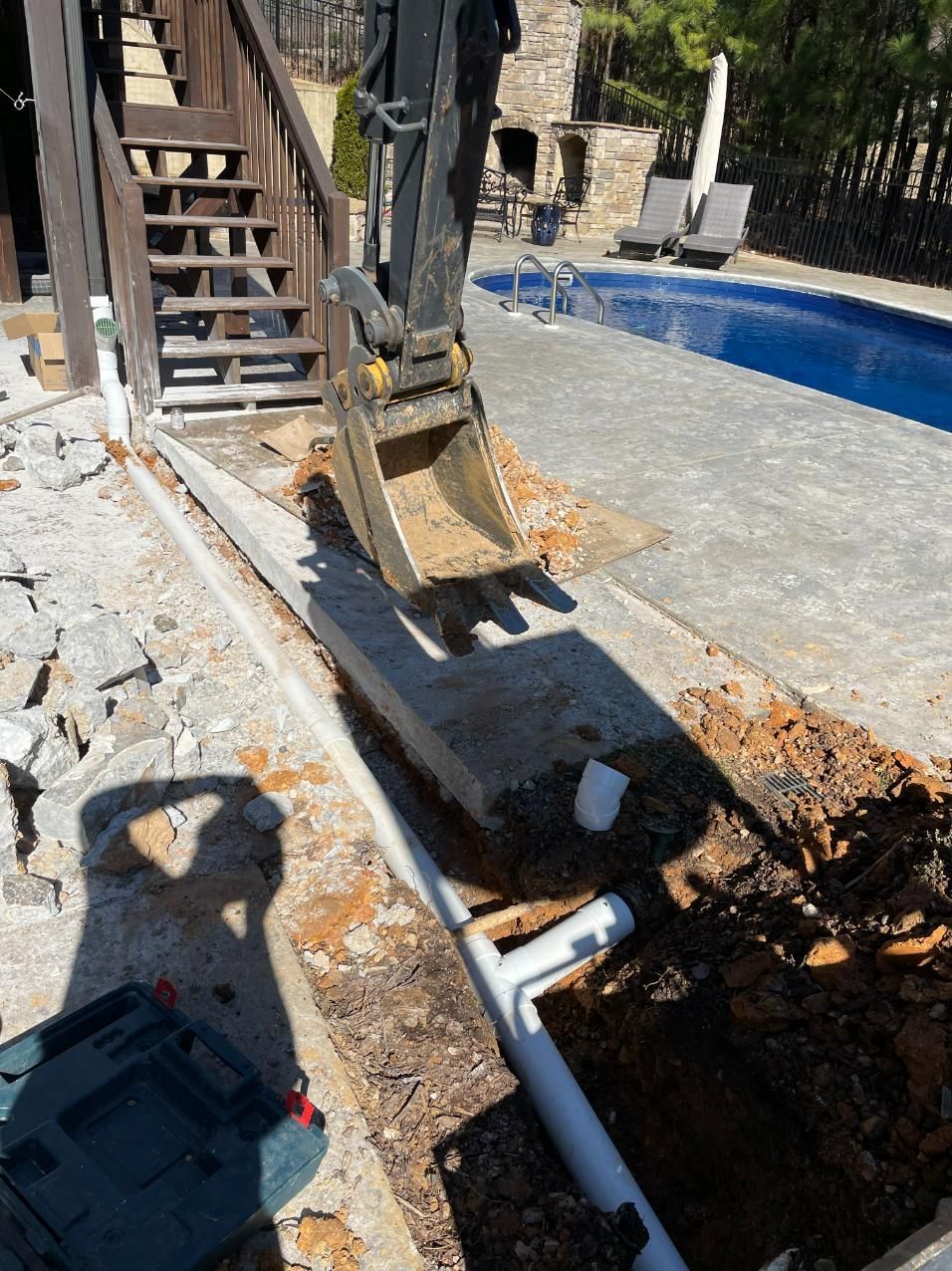 An excavator digging near a swimming pool. Brown soil, white pipes, and a gray concrete patio are visible.