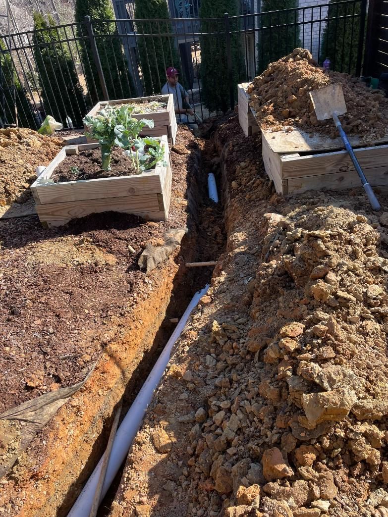 A trench dug near raised garden beds, with white PVC pipe installed. Brown dirt piles and a shovel are present.