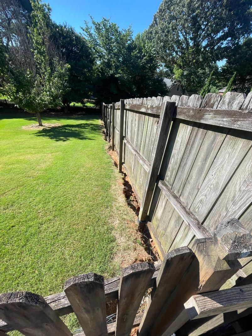 Wooden fence bordering a yard with green grass and trees under a blue sky.