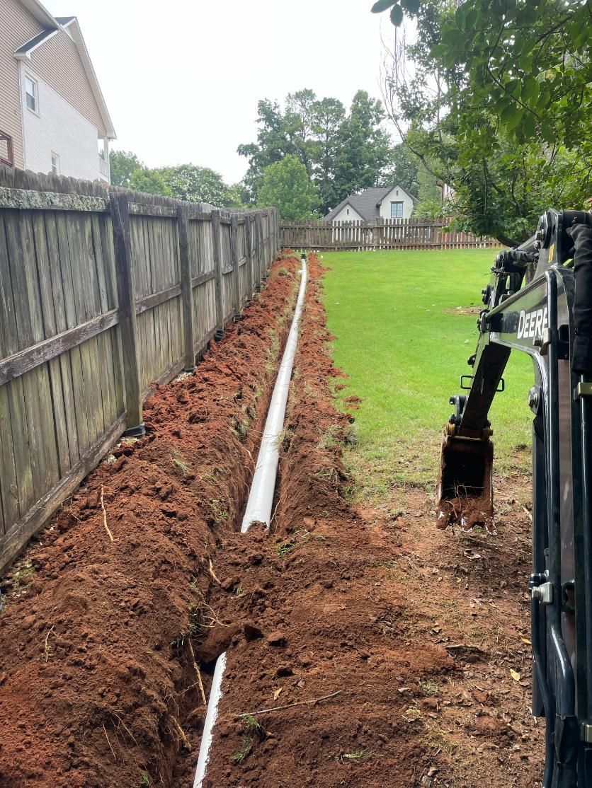 A trench along a fence with white pipes installed. Mini excavator on the right.