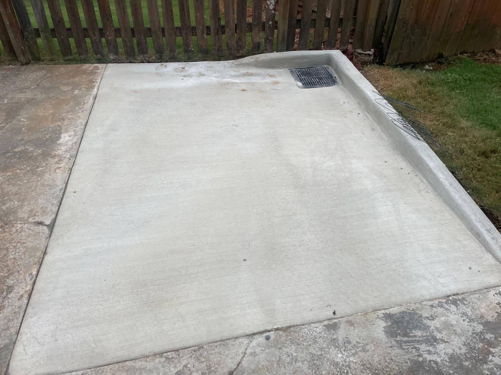 Concrete patio with a dark grate, next to grass and a wooden fence.