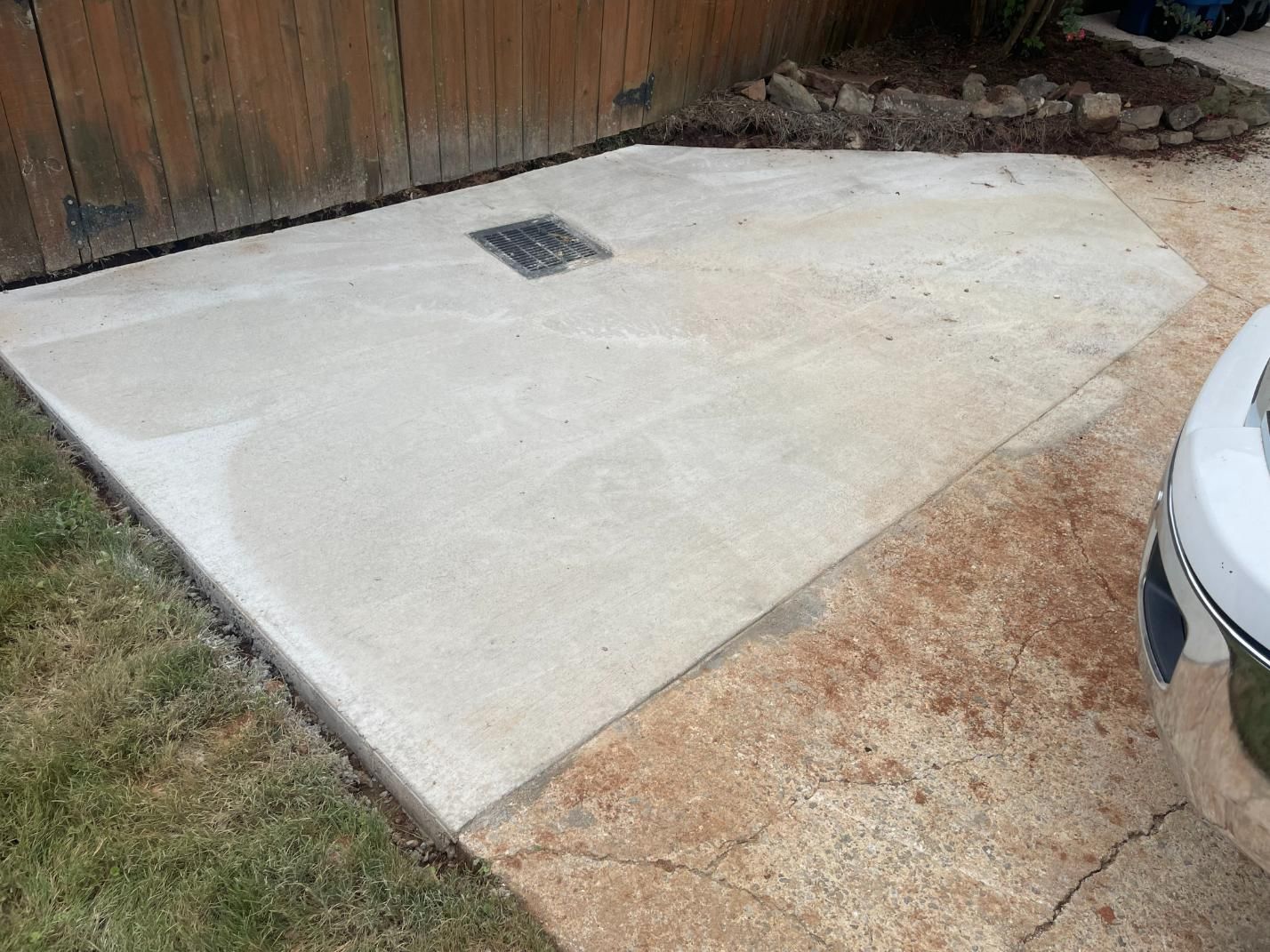 Concrete patio next to a wooden fence and driveway; a drainage grate is visible.