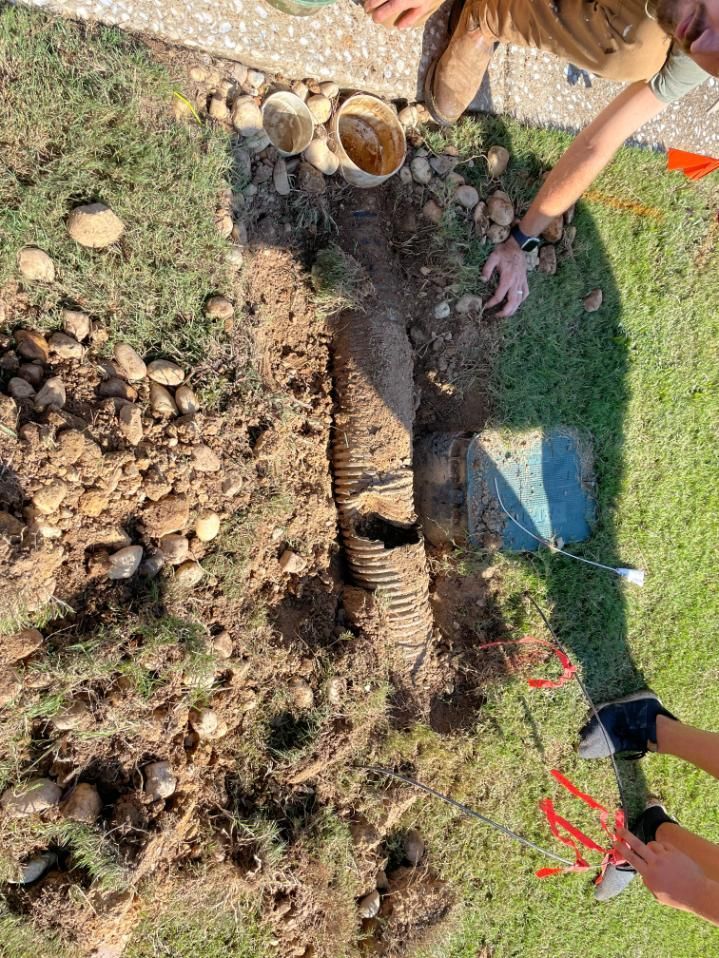 People working on a drainage system in a grassy area; corrugated pipe and dirt visible.