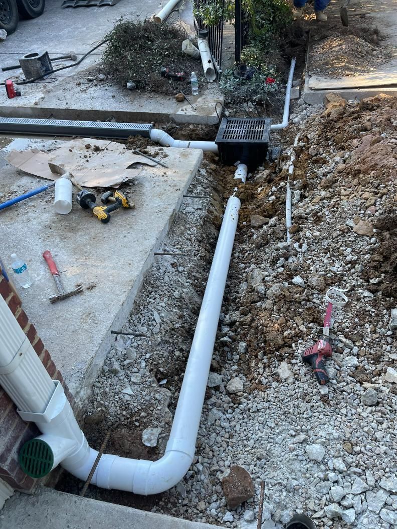 White pipes being installed in a trench next to a concrete walkway and a brick building, with tools and dirt visible.