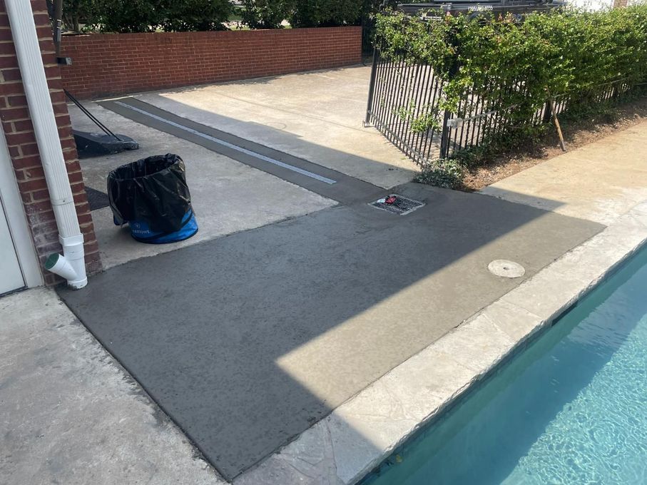 Concrete patio next to a swimming pool, brick wall, trash can, and a drain.