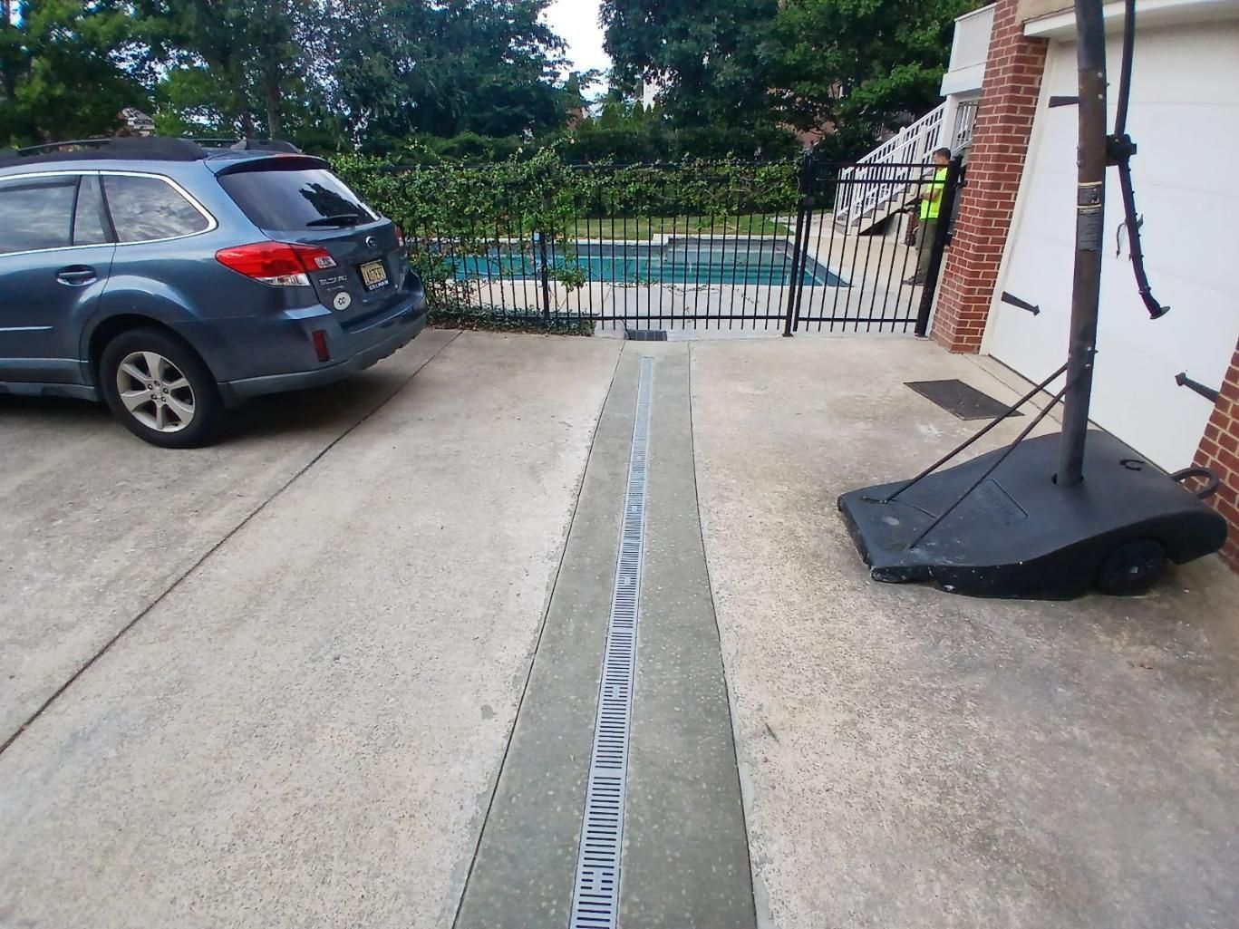 Blue car parked near a long concrete drain in a driveway. A pool is visible behind a fence.