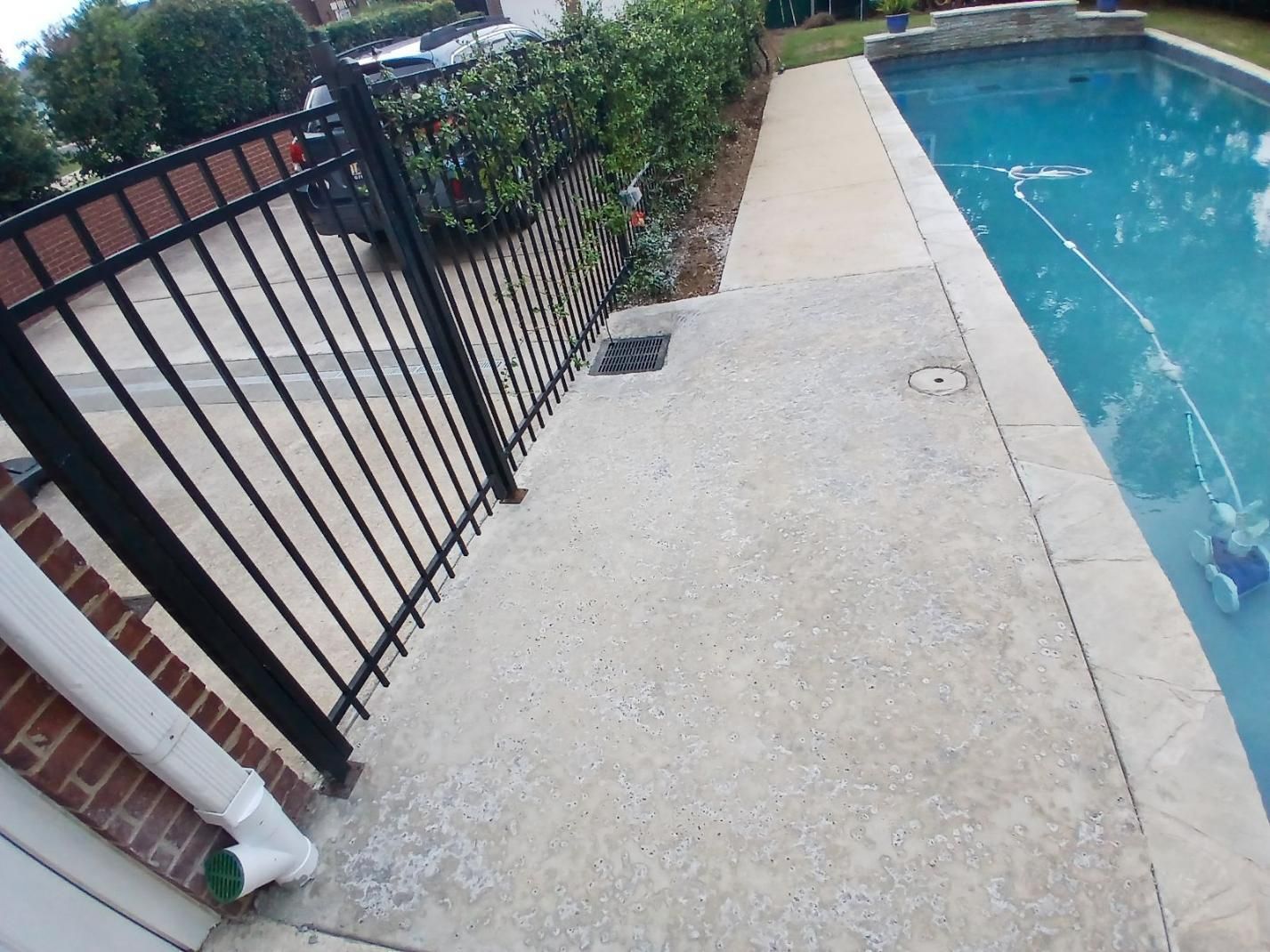 Poolside with concrete walkway, black fence, and bushes. Water is in the pool, with a brick building on the side.