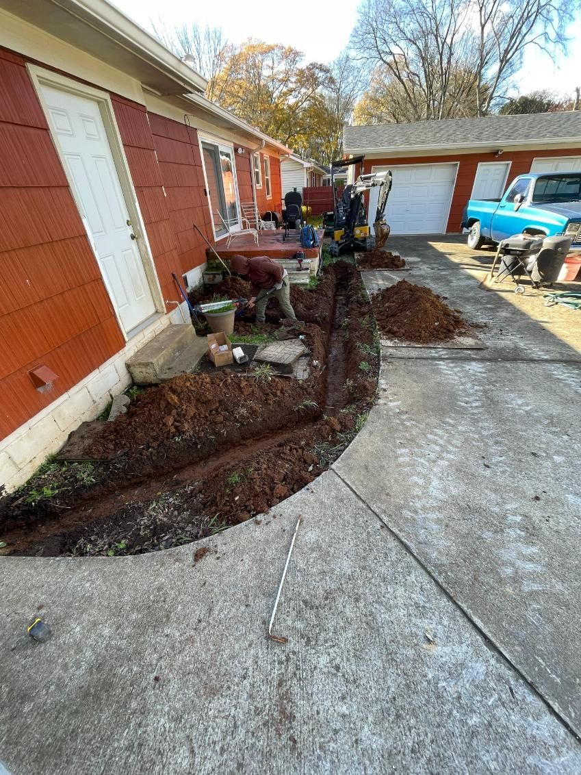 Workers digging a trench along a red building's side, moving dirt onto a concrete driveway.