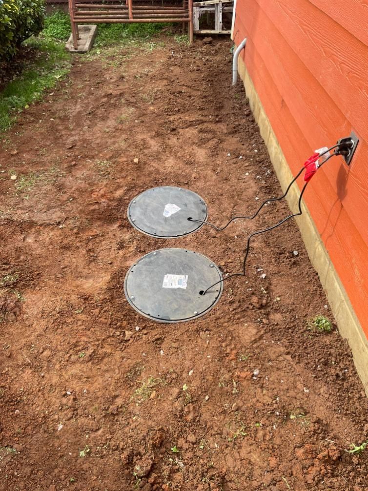 Two circular gray lids on the ground near an orange building. Wires run from them to the building. Brown dirt.