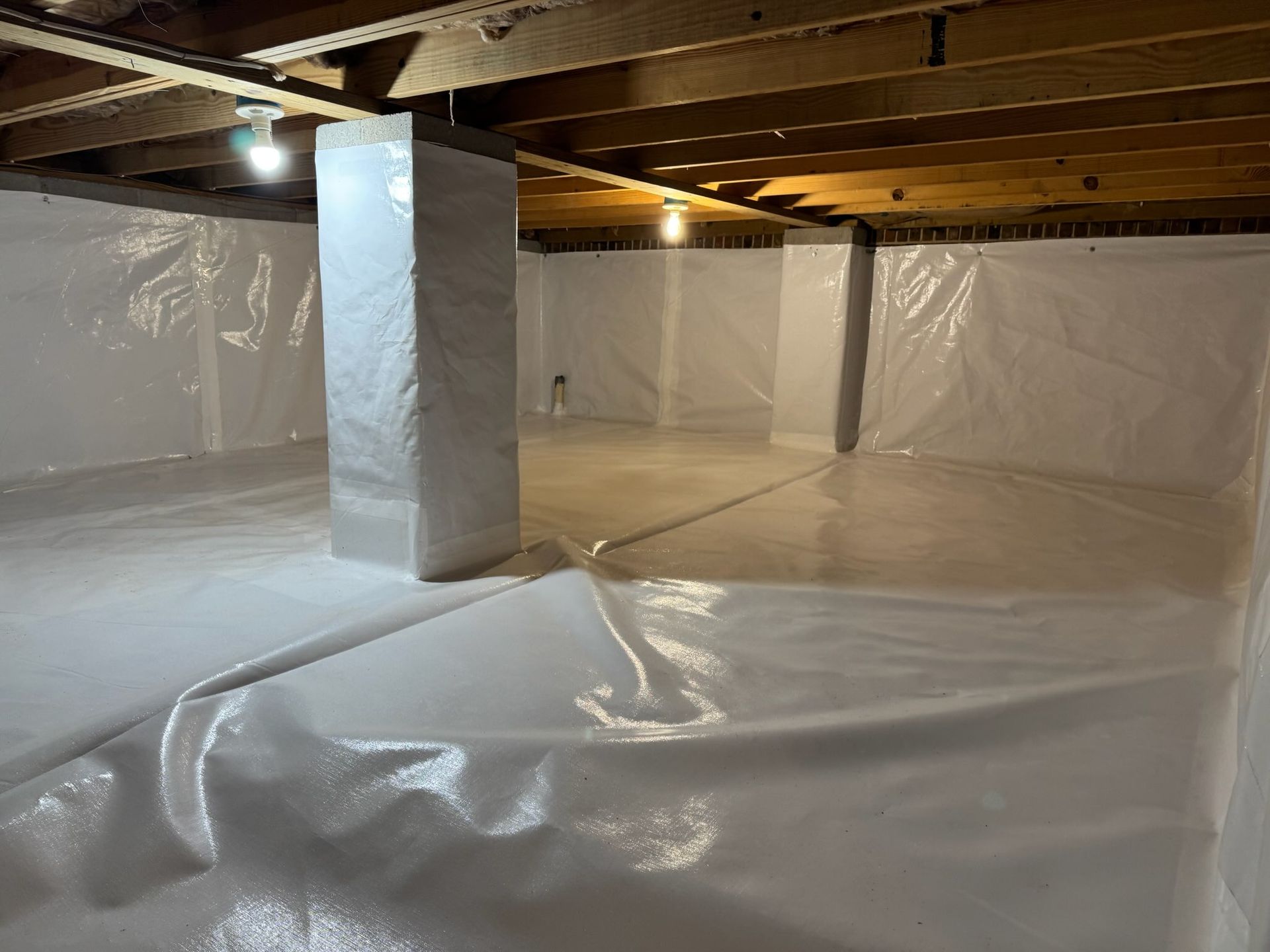 White-walled crawlspace with white plastic on the floor and support pillars, lit by overhead lights.