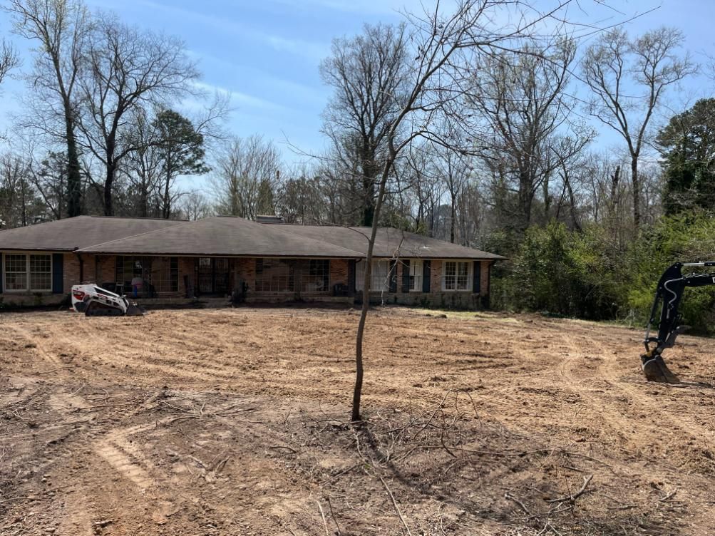 House with excavated yard, surrounded by trees. An excavator sits to the right.