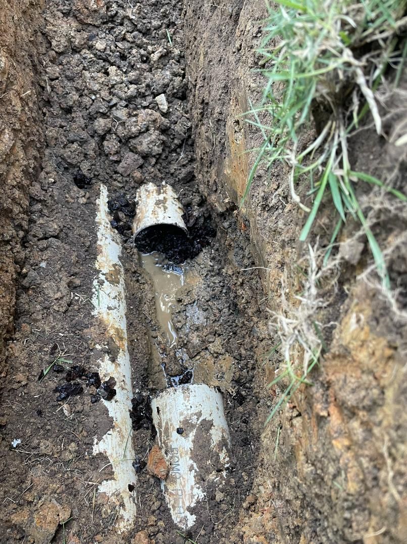 A trench with exposed white PVC pipes, one cracked, covered in dirt and oil. Grass is seen on the side.