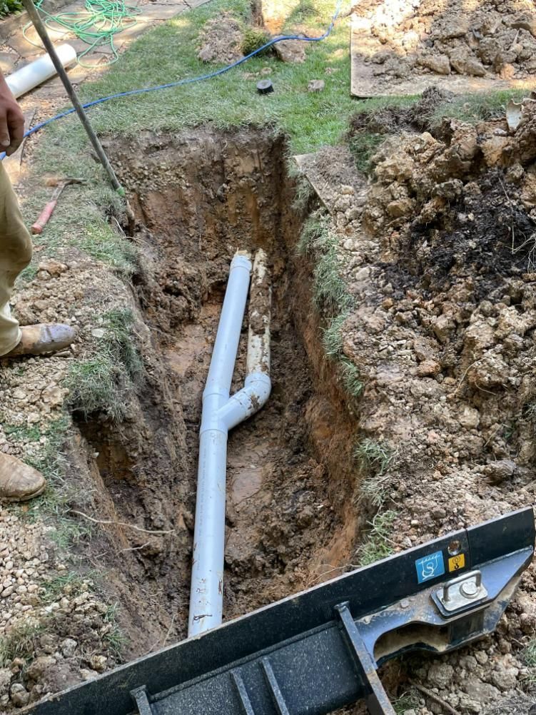 Trench with plumbing pipes installed, person holding a rod. Outdoor setting with soil and grass.