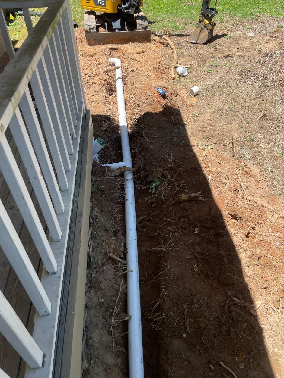 Trench dug beside a house, with white PVC pipe installed. Small excavator in background. Sunny outdoors.