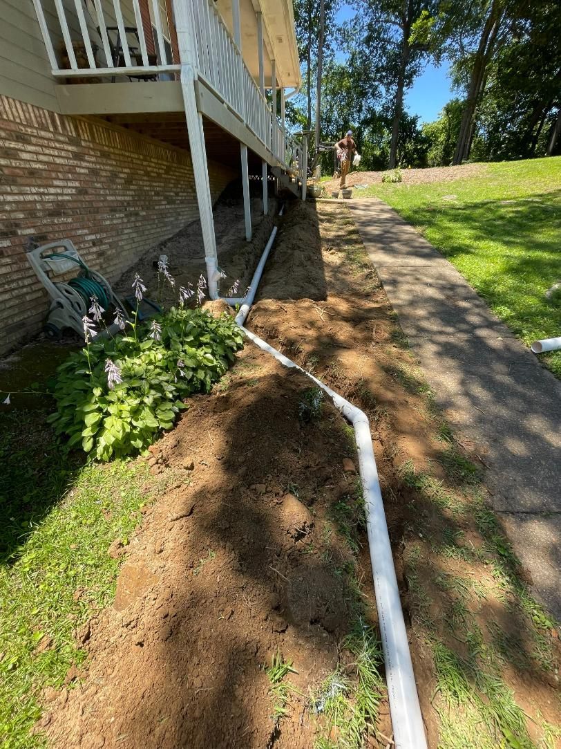 A person installs white drainage pipes near a brick house and a walkway, outdoors.