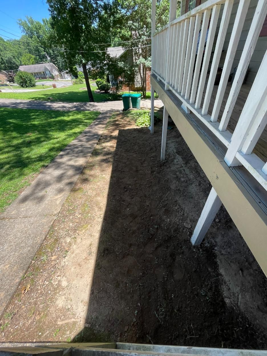 A long, narrow dirt path next to a porch with white railings and posts. Green bins are in the distance.