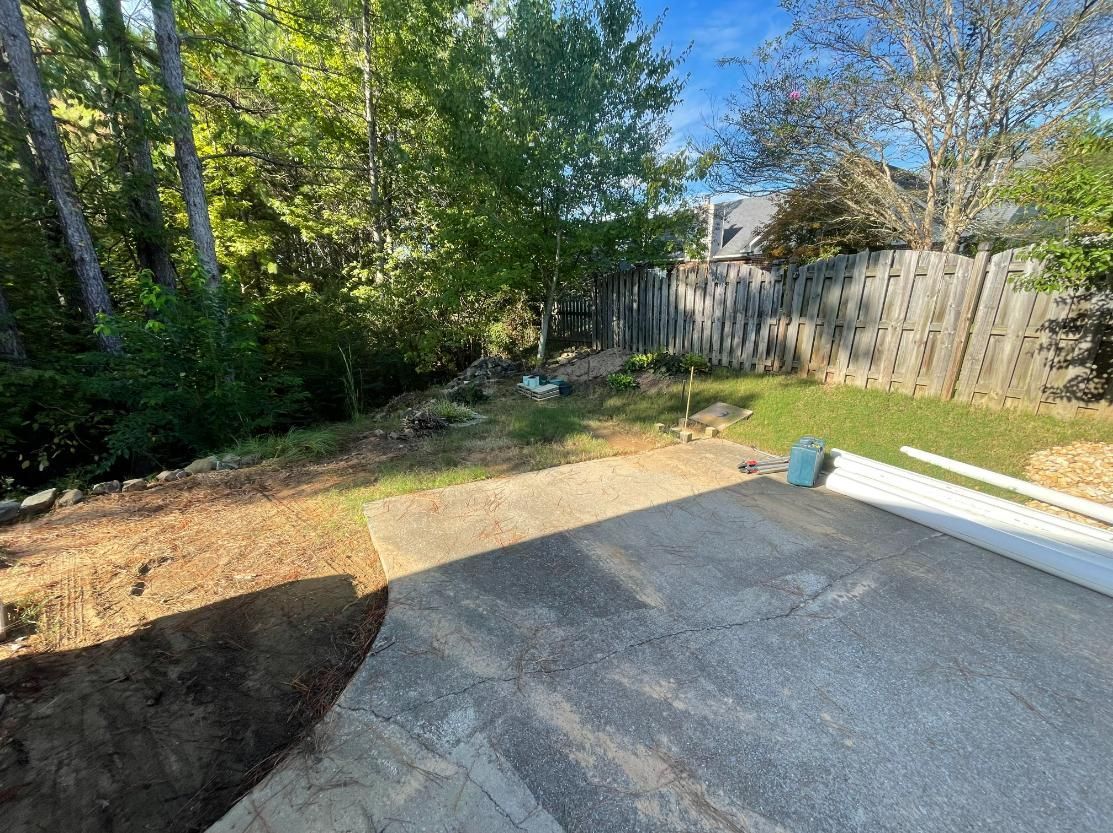 A backyard with a concrete patio, grassy area, trees, and a wooden fence.