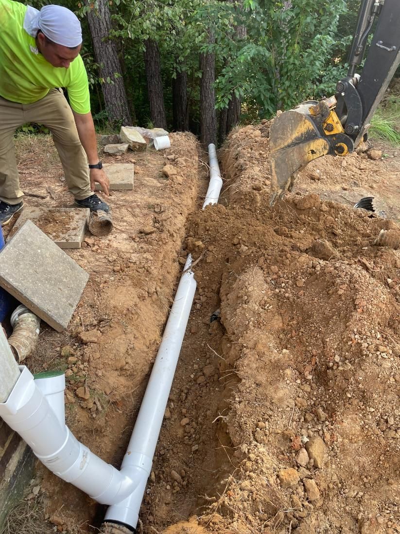 Man installing white PVC pipes in a trench outdoors, near trees, with backhoe in background.
