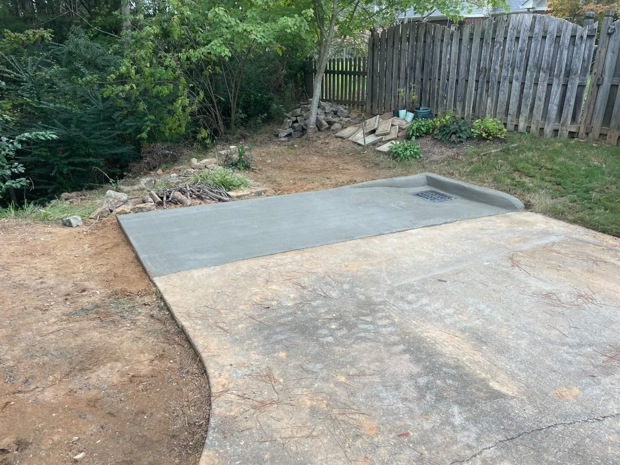 A new concrete slab next to an older one, in a yard, with a wooden fence and greenery in the background.